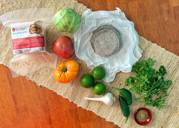 All the ingredients you need for this recipes laid out on a table: Bartleby's Spicy Crispy Cutlets, green cabbage, red and orange tomatoes, blue corn torillas, three limes, a head of garlic, jalapenos, fresh cilantro and parsley, and a small bowl with oregano.