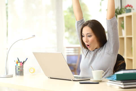 woman at work in front of laptop looking happy with arms raised