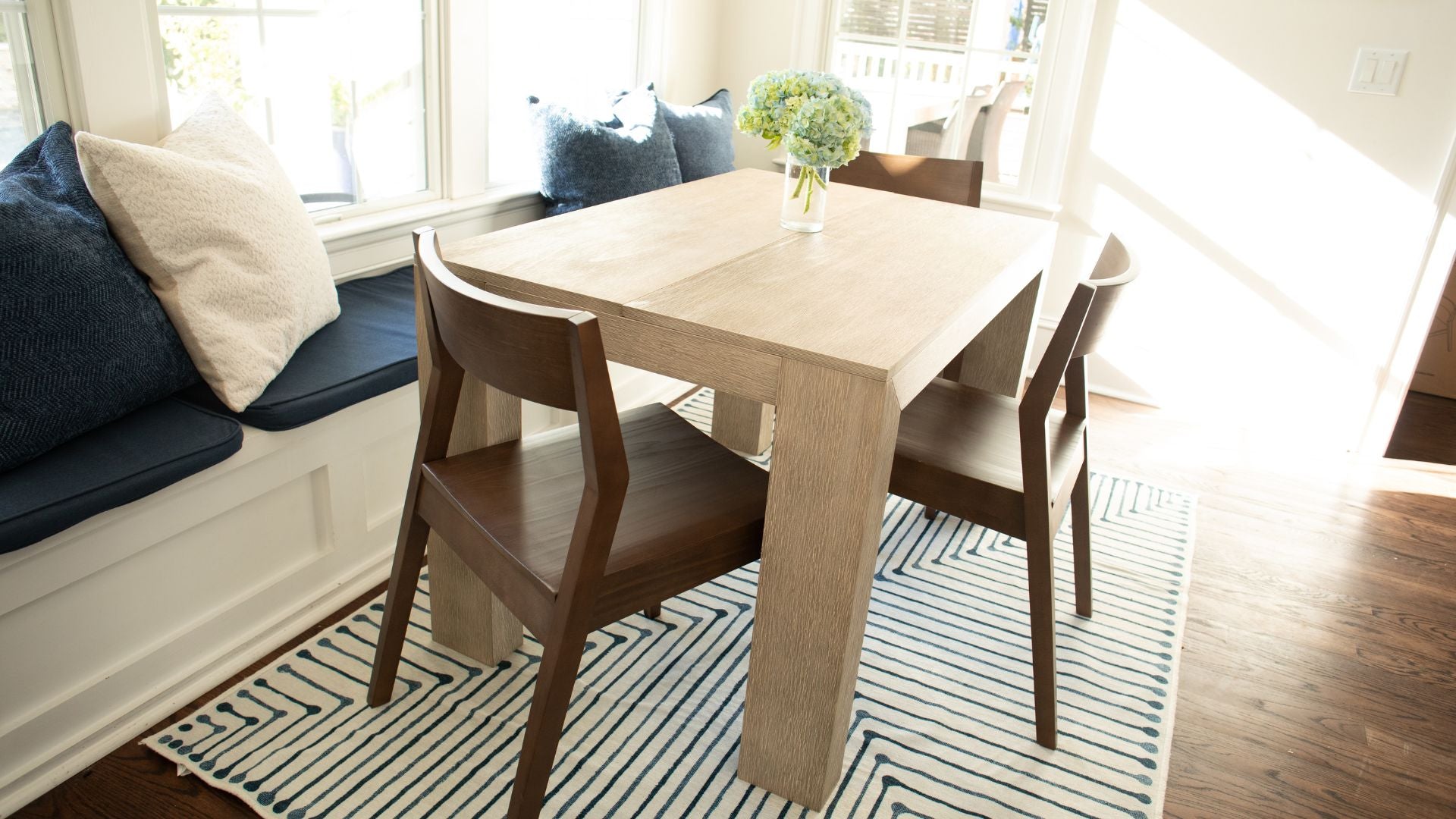 Solid wood modern dining table in seashell with solid wood dining chairs in walnut, banquette, striped rug, and floral centerpiece