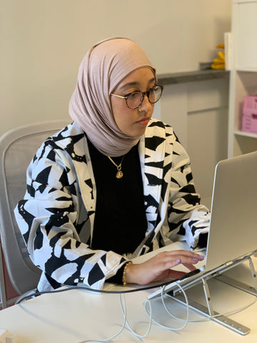A muslim woman in a headscarf sits at her desk working on her computer, wearing black & white patterned pyjama jacket 
