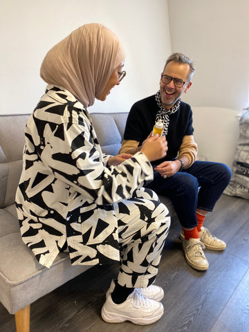 Muslim woman in headscarf and monochrome pyjama suit on sofa with white man wearing glasses have a conversation 