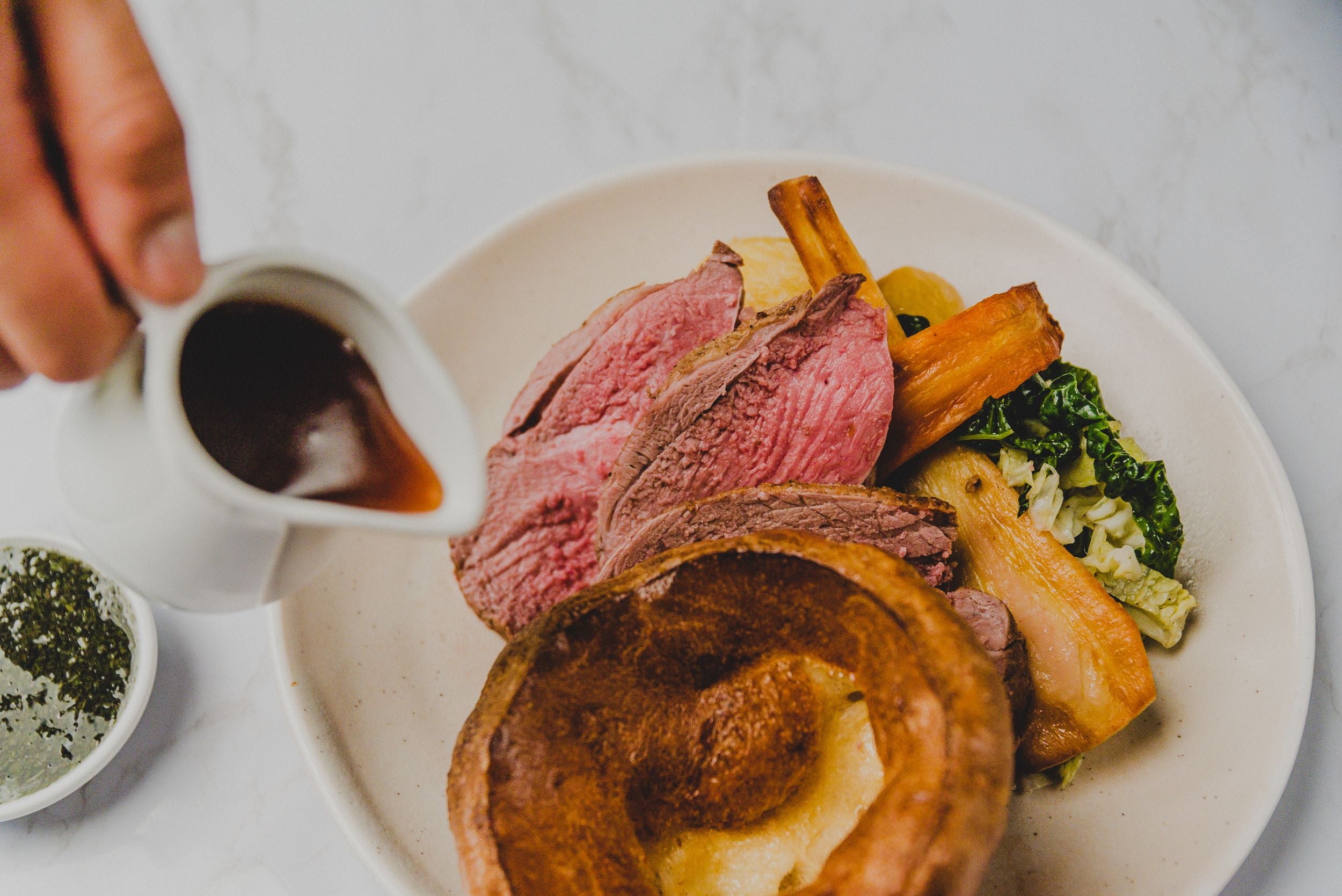 Roast beef, yorkshire pudding and vegetables on a plate. Someone pours gravy over the top. 