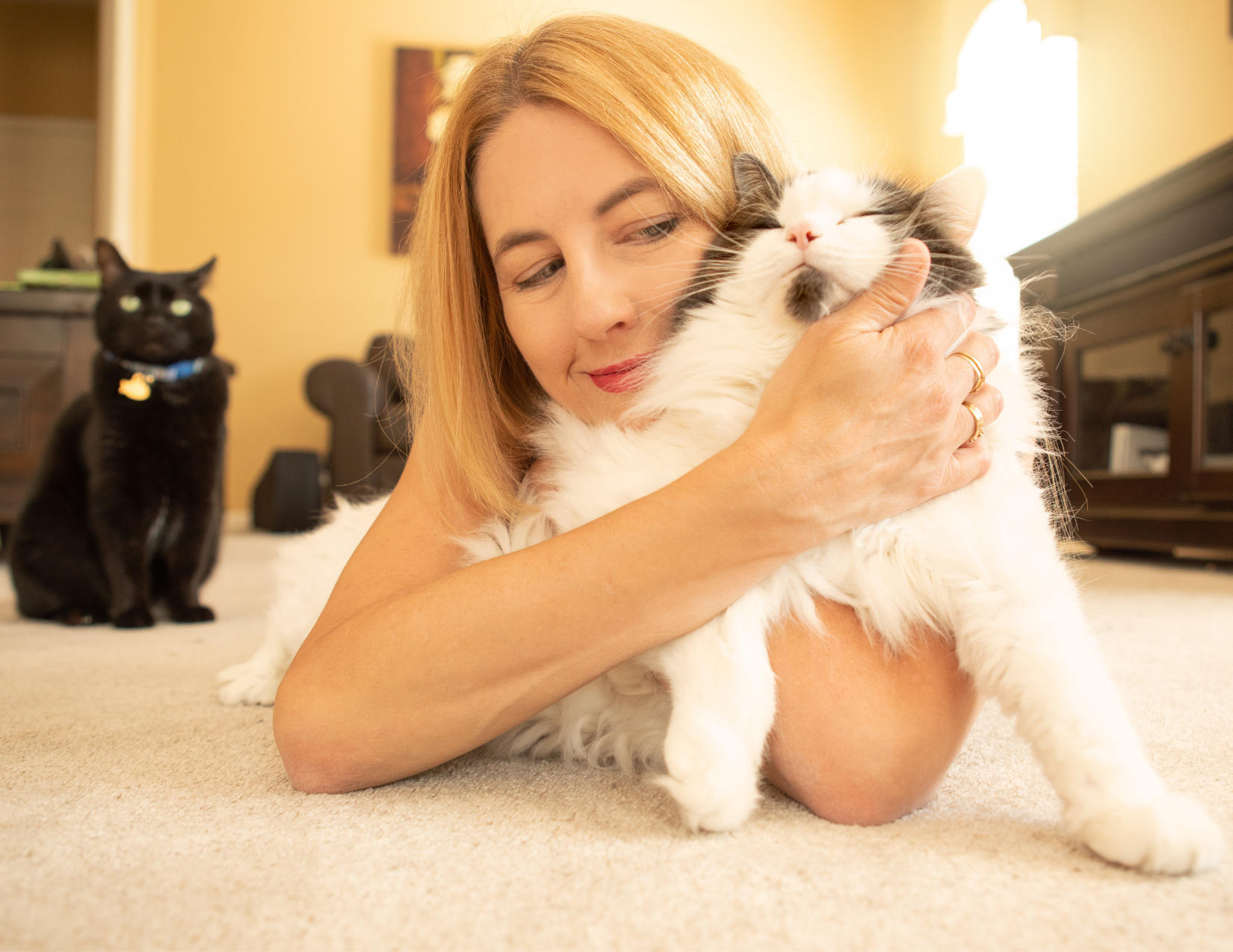 Woman cuddles her cat and while her other cat waits for their turn.