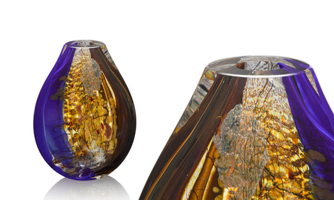 A sculptural blown glass vessel by Vermont glassblower Randi Solin features layers of deep blue glass, silver foil, warm golden glass, and chocolate brown glass against a white background.