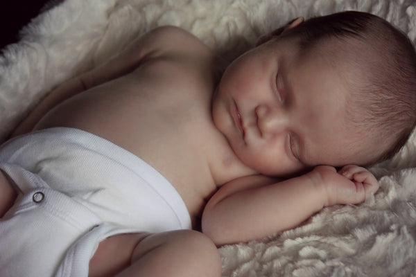 Baby som ligger på ryggen på et offwhite teppe og sover med hodet hvilende mot armen sin.