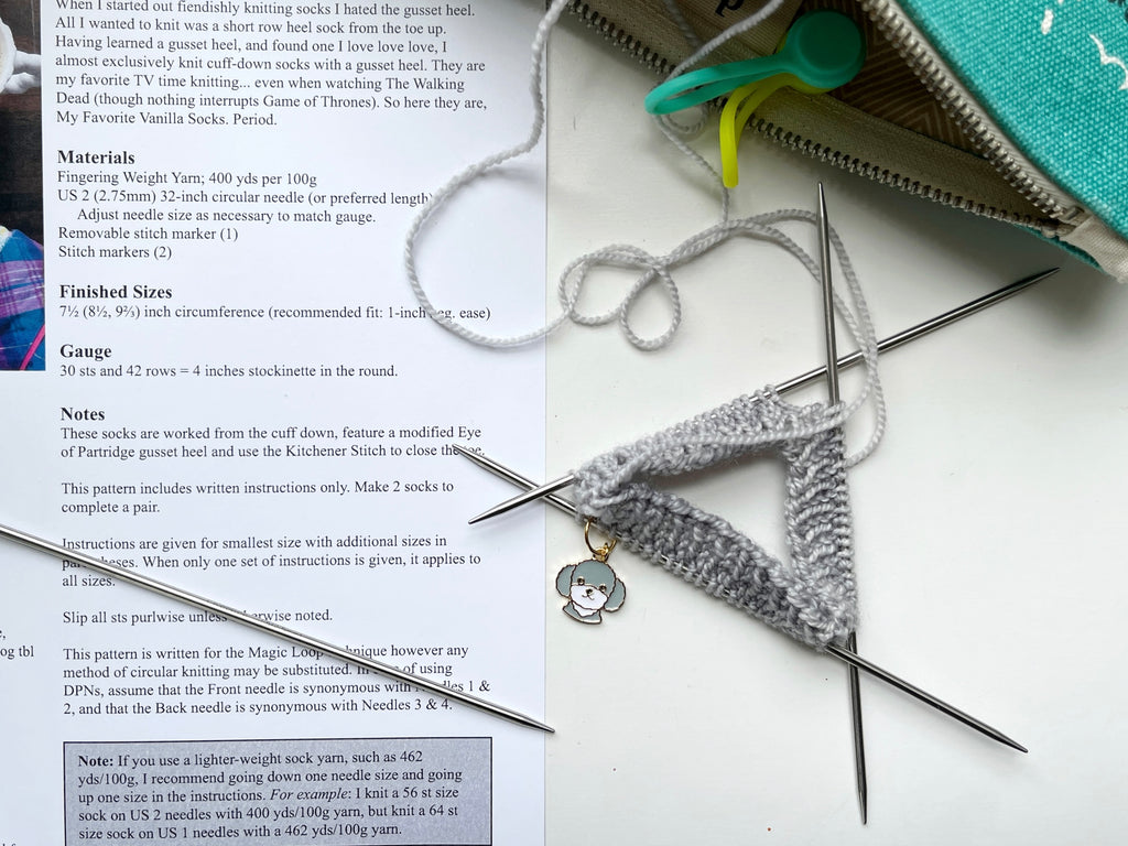 A grey doodle dog beginning of the row stitch marker in use on an in-progress sock knitting project, being knit on a set of double point needles. 