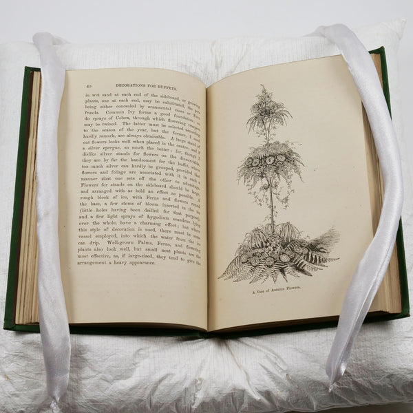 Image of a book open on a cushion. The right-hand page is an engraving of a large and elaborate, three-tiered floral arrangement including ferns. The left-hand page is text.