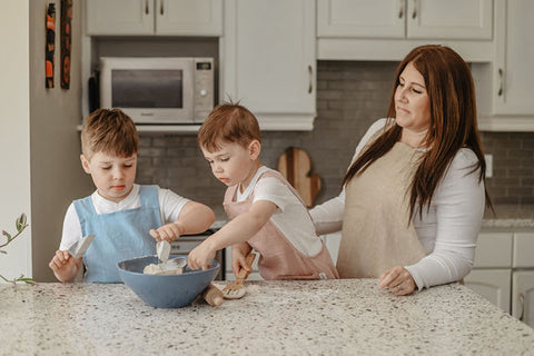 Two boys in aprons in a kitchen with a mixing bowl while their mom watches