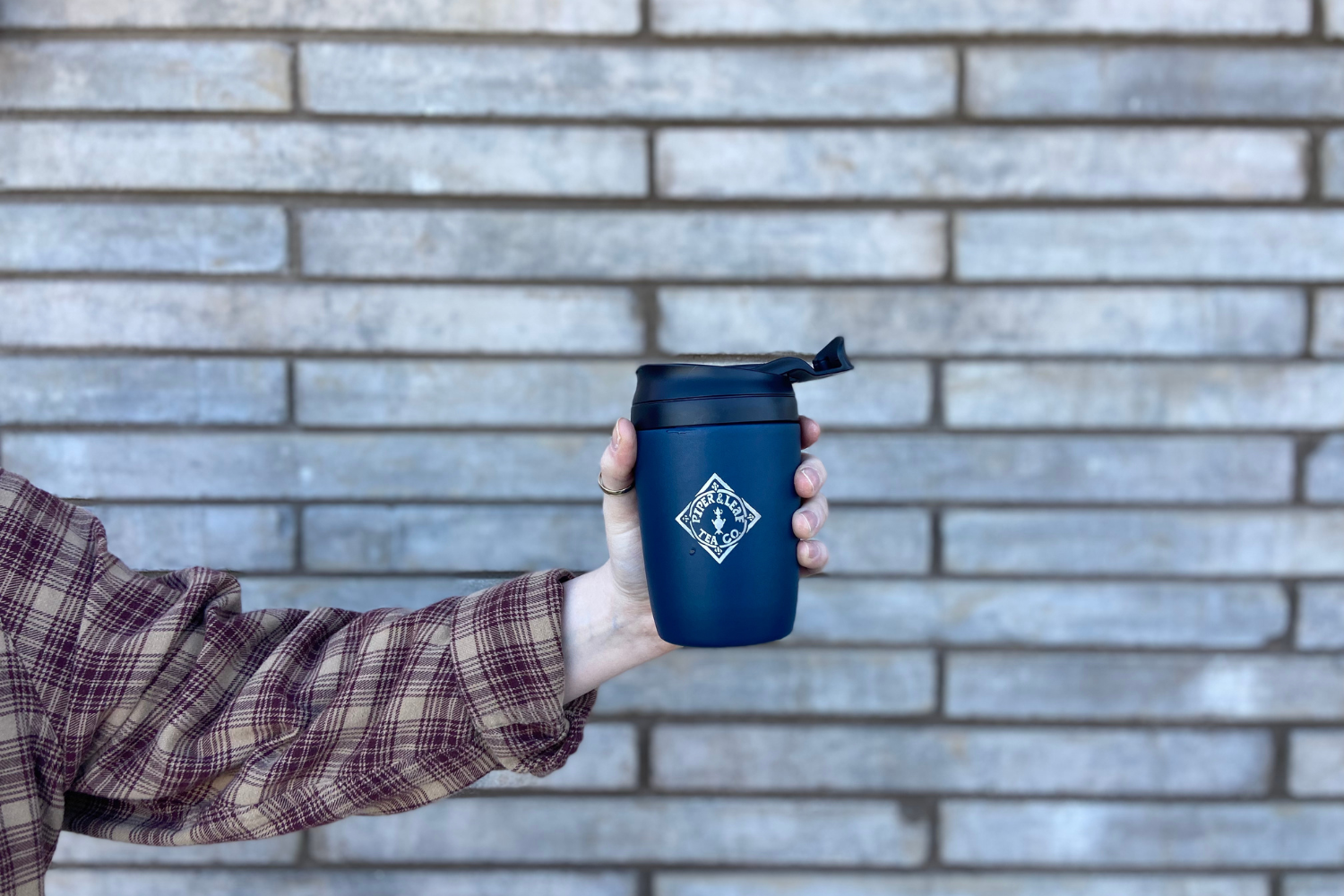 A hand holding a Piper and Leaf insulated travel mug in front of a brick wall
