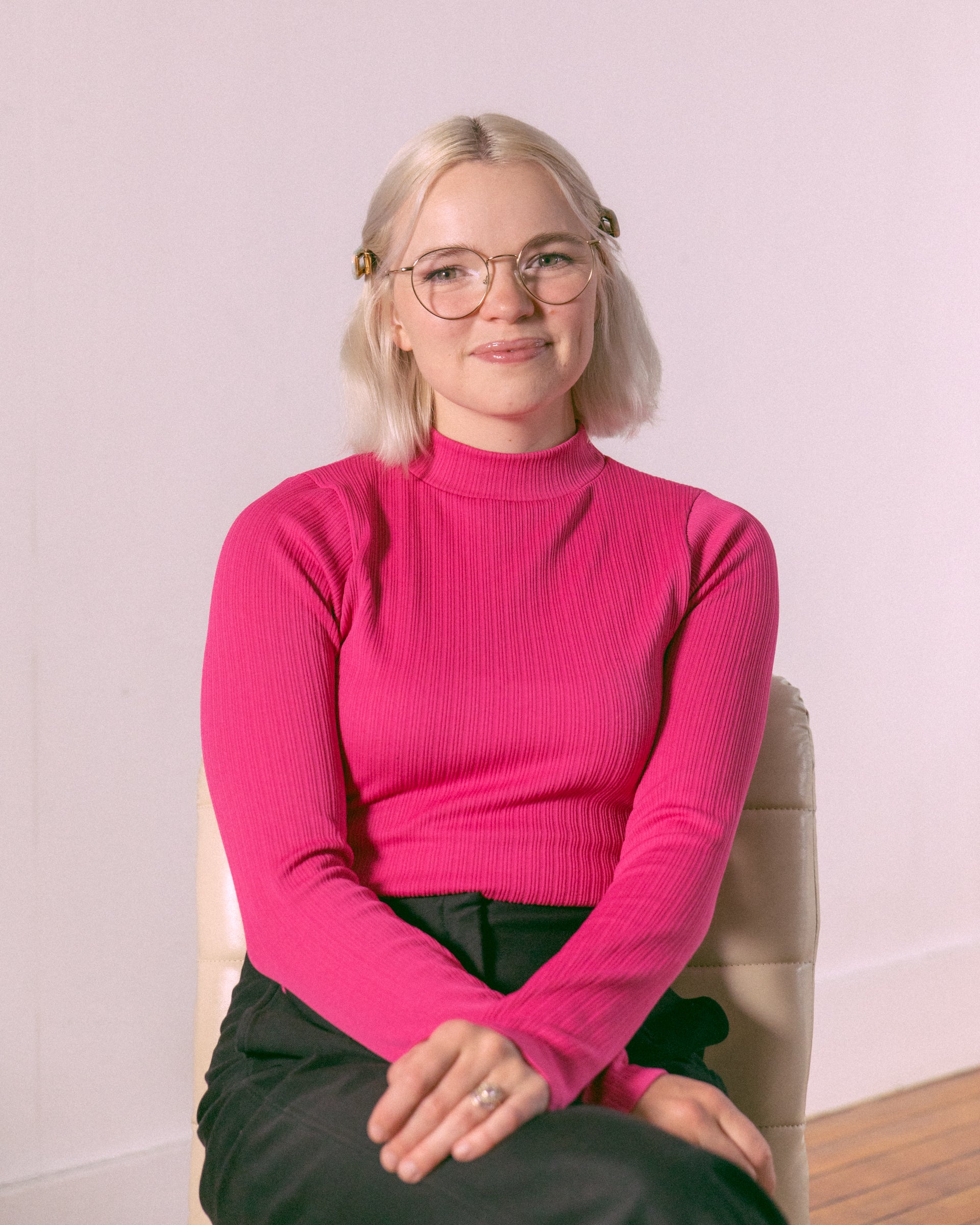 Georgia's presence radiates against the white walls as she sits comfortably in a beige chair. The soft, natural light from the window perfectly complements her pink turtleneck, creating a warm and inviting atmosphere. Bertie, as the photographer, skillfully captures the subtle nuances of Georgia's personality, making each shot feel intimate and genuine.