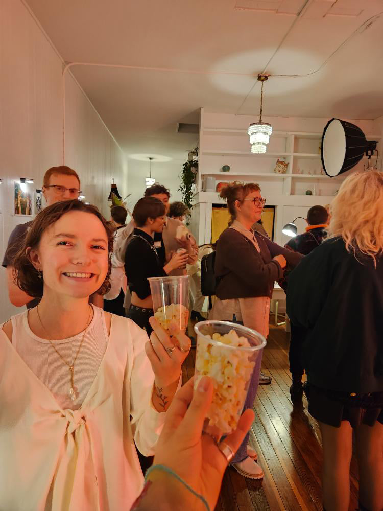 A crowd of people are walking and talking in the studio, one person is smiling at the camera and holding a cup of popcorn. Another hand holding a cup of popcorn is in the bottom center of the image.