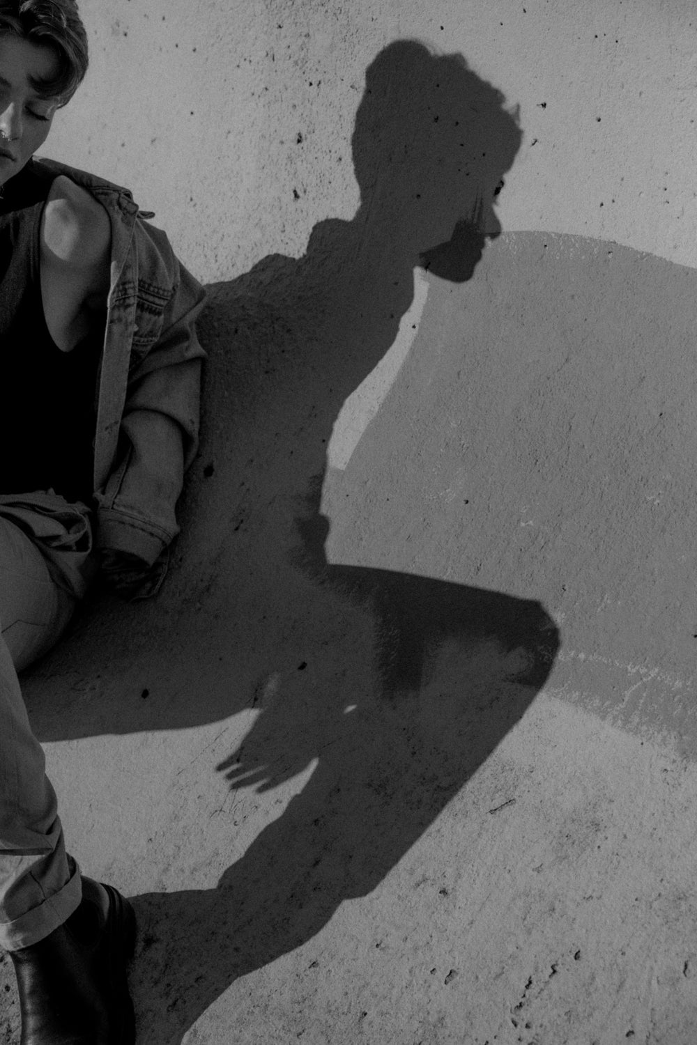 Non binary model leaning against grungy cement wall. The model is cropped and the focus of the image is on the shadow. They are wearing a jean jacket and have short hair.