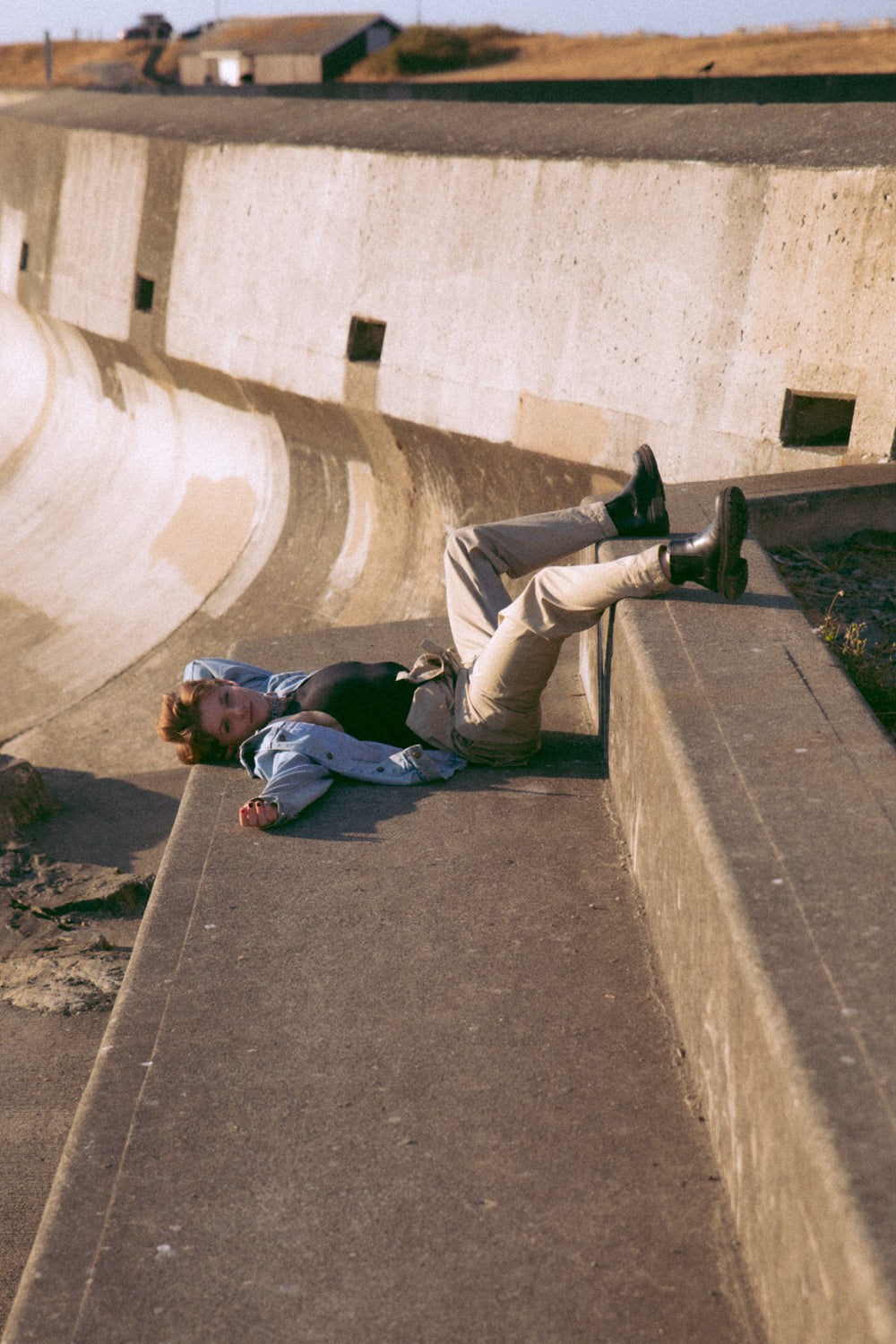 Non binary model sitting upside down on some large cement stairs. They are wearing a jean jacket and have short hair.