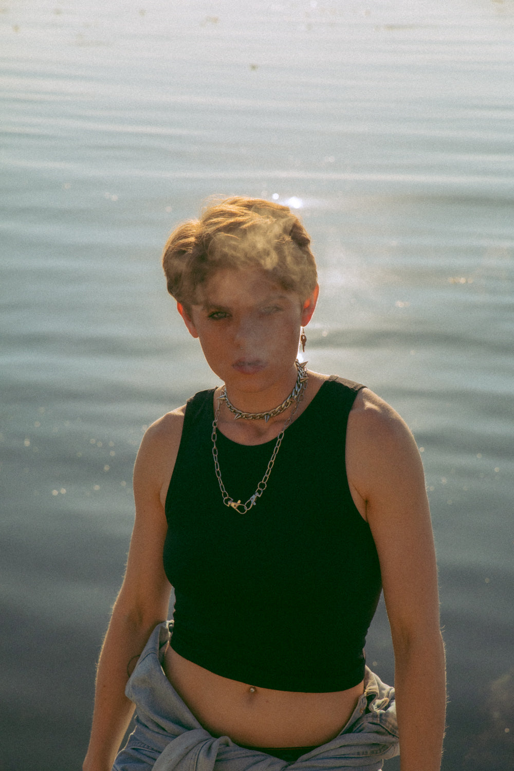 Non binary model exhaling smoke. The smoke is partly covering their face while they stare at the camera. They are wearing a black tank top, one key-shaped earring and two chain necklaces (one with spikes). The ocean is behind them.