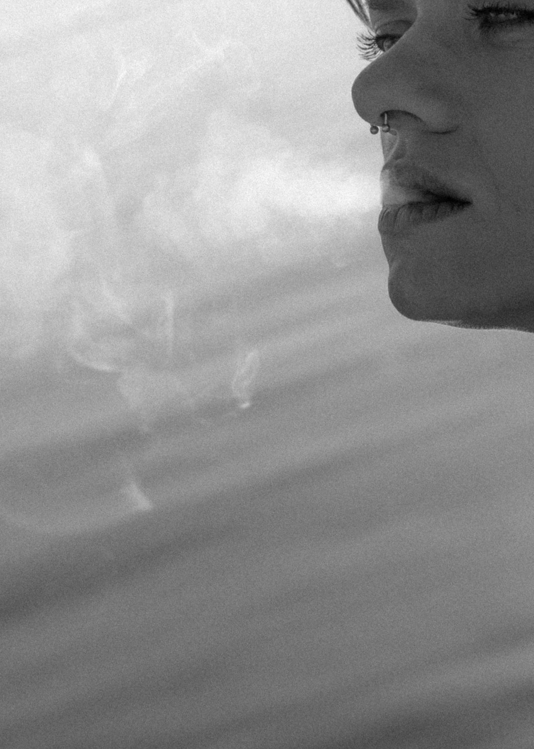 Non binary model exhaling smoke. The ocean is behind them. The photo is cropped so we only see their face in the top right corner. They are looking toward the left. Their body, ears, forehead and neck are cropped out.