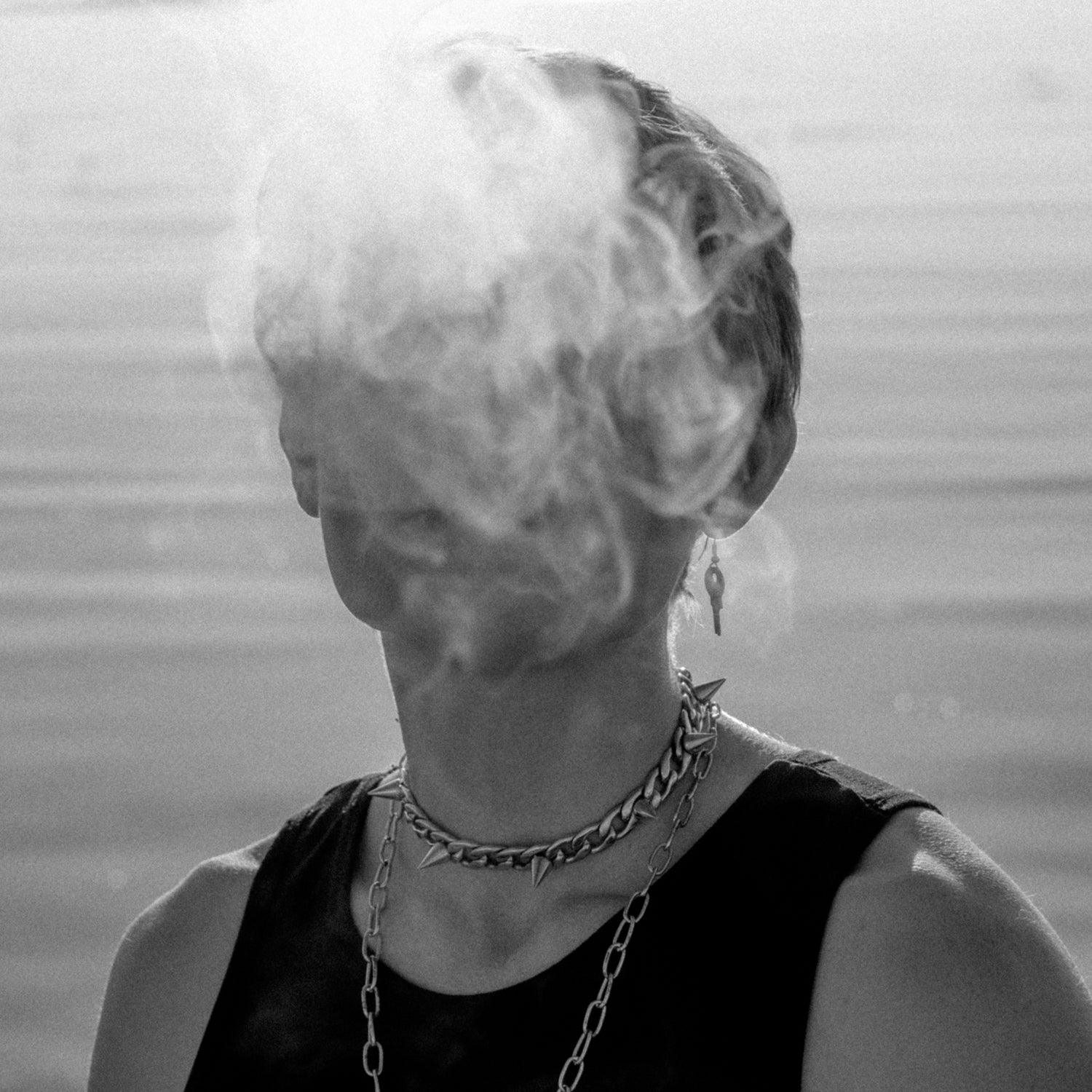 Non binary model exhaling smoke. The smoke is covering their face. They are wearing a black tank top, one key-shaped earring and two chain necklaces (one with spikes). The ocean is behind them.