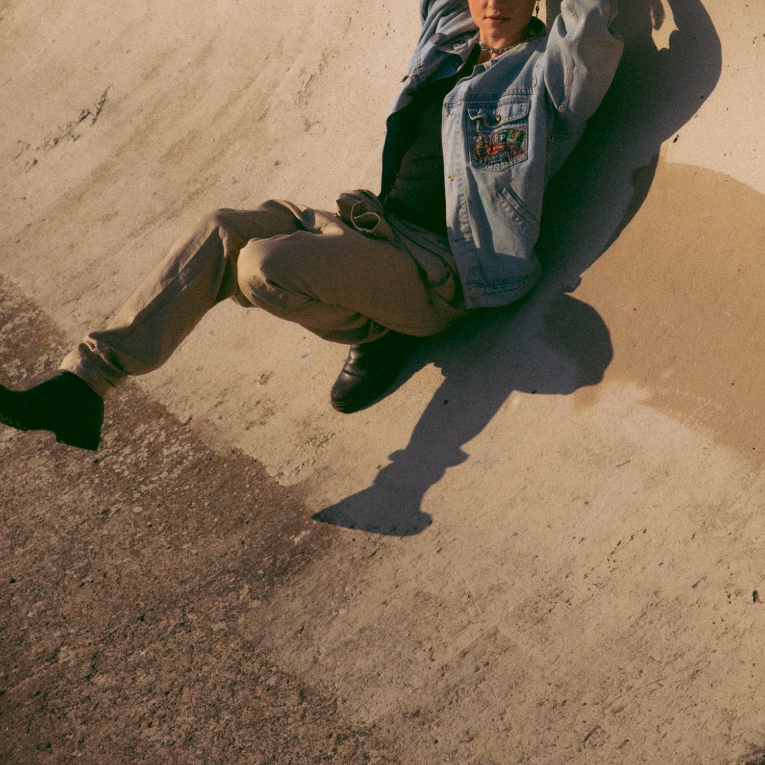 Non binary model leaning on grungy cement wall with one leg in the air. They are wearing a jean jacket and have short hair.