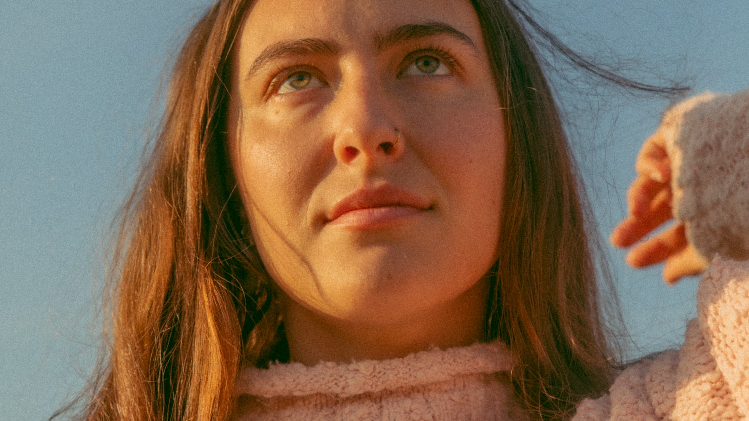 Close Up Face shot of model Lau who is looking upward into the distance, while brushing her right hand through her hair. She is wearing a pink knitted turtle-neck sweater and the background is blue sky.