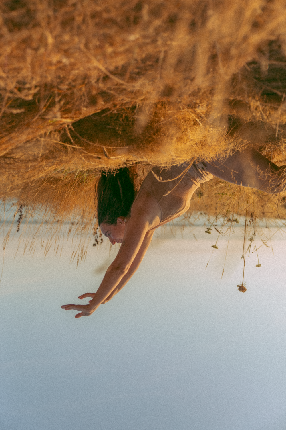 Model is sitting in a field with arms reaching up toward the sky. Tall yellow grass and blue skies make up the surroundings. We see the ocean in the distance. The image is flipped vertically so the model appears to be upside down. Lau is wearing a beige slip.