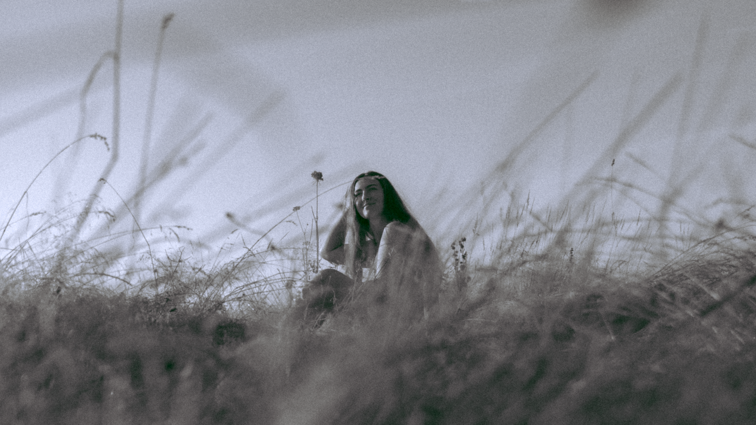 Model is sitting in a field smilling and resting her head on her right hand. She is centered in the shot and tall grass is blurred in the foreground. Black and white.