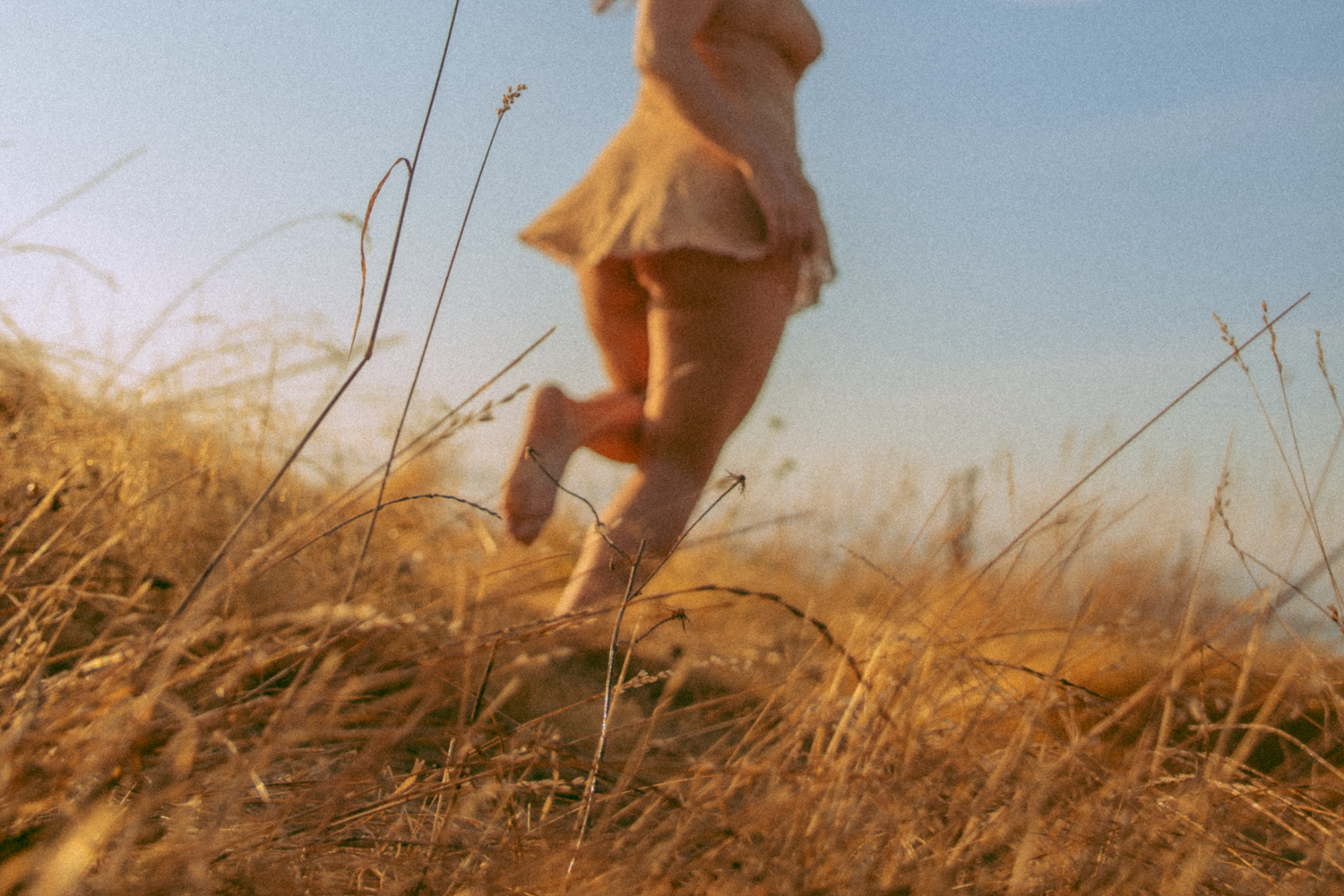Model is running away from the camera in a field, her head is cut off. Tall yellow grass is in focus in the foreground, blue sky in the background. Lau is wearing a beige slip.