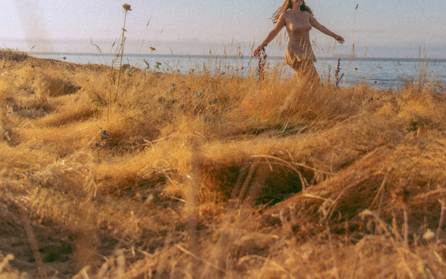 Model is dancing in a field with arms out, her face is cut off above her smile. Tall yellow grass and blue skies make up the surroundings. We see the ocean in the distance. Lau is wearing a beige slip.