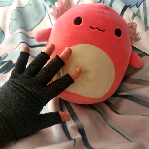 Sharon's hand in a grey compression glove, on top of their Archie the Axolotl Squishmallow