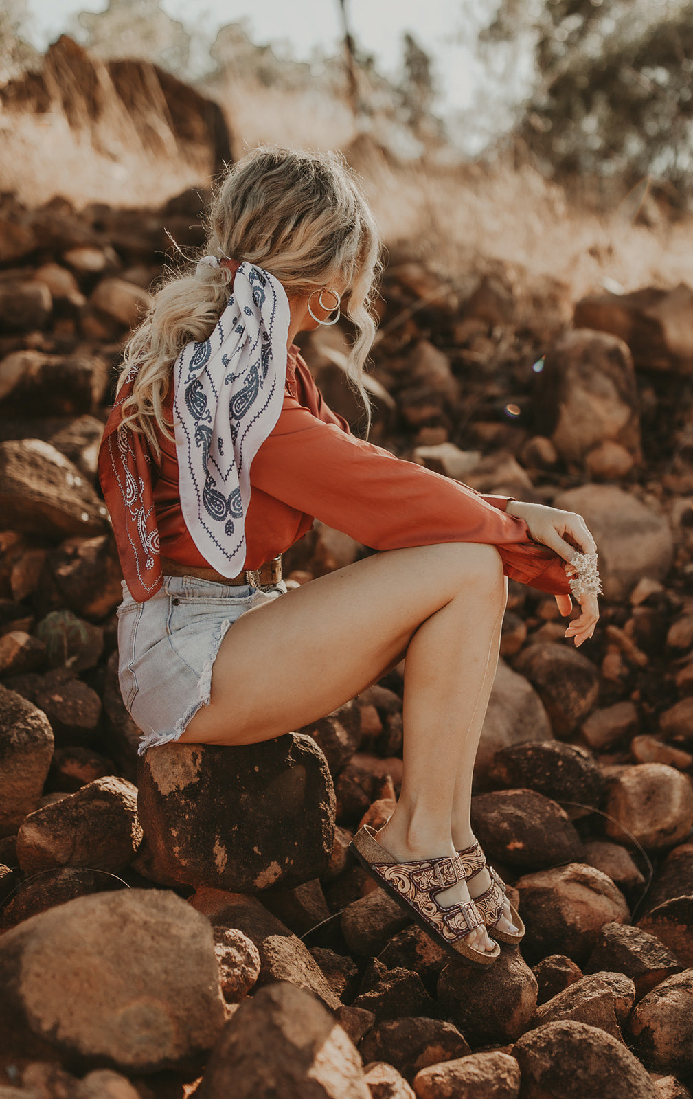 A blonde haired model sits on a riverbed of stone. She wears a red top, denim skirt and Branded Faith shoes.