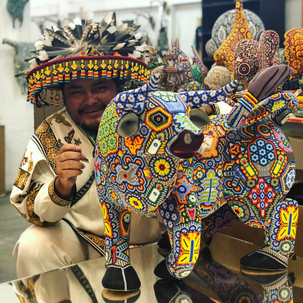 Artista huichol vistiendo sombrero con plumas y traje tradicional, elaborando una pieza de arte que es un toro multicolor.