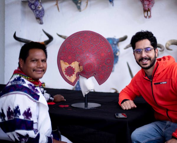 Un artista huichol y Fernando Motilla con una pieza de arte escultura de Cesar Orrico intervenida con arte huichol en color rojo.
