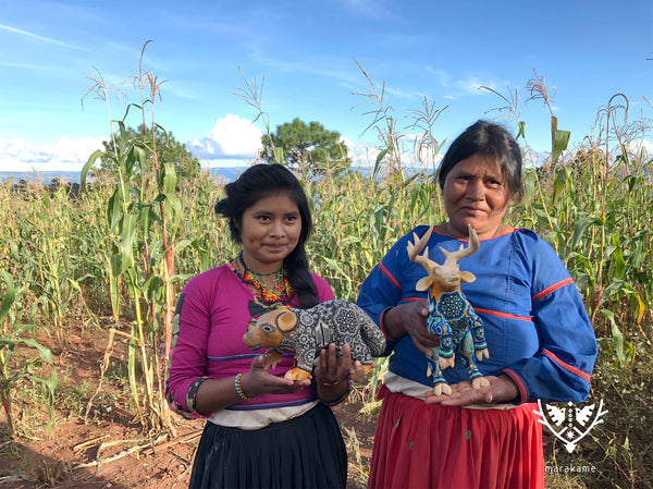 Dos mujeres huicholes con dos esculturas, un oso y un venado en colores gris y azul, se encuentran en un sembradío de maíz.
