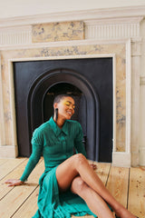Light brown skinned woman sits with legs crossed on wooden floor in green dress.