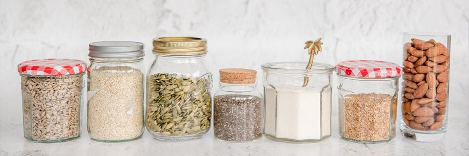 Row of 7 glass jars containing the whole food ingredients used to make Purition; sunflower kernels, sesame seeds, pumpkin seeds, chia seeds, coconut, flaxseed & almonds.