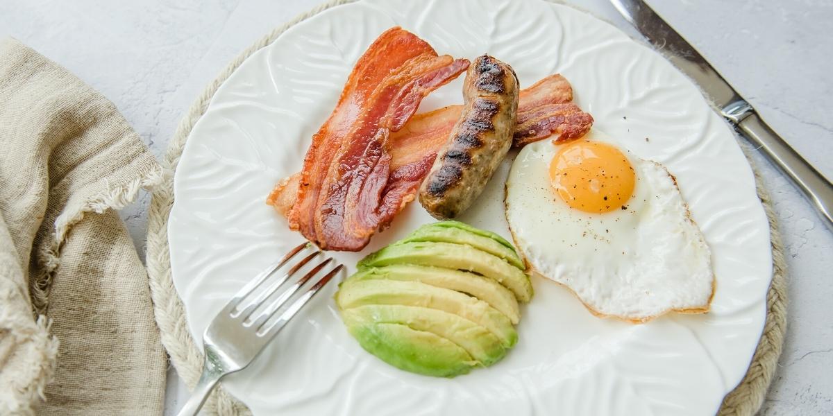 English breakfast on a plate; bacon, sausage, fried egg and half an avocado.