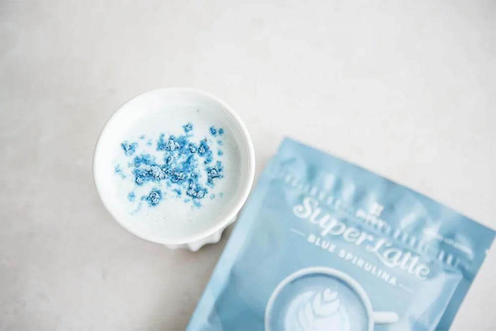 A pack of Purition's Blue Spirulina Super Latte next to a white porcelain cup of Blue Spirulina Latte.