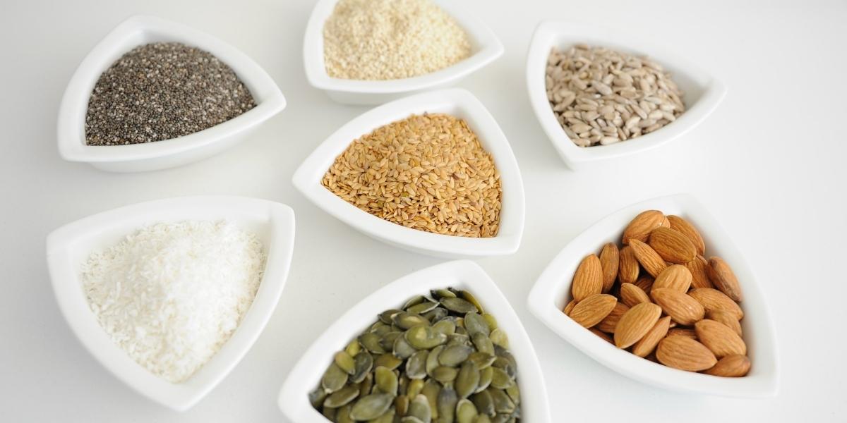 A photograph of the 7 whole foods used in all Puriton wholefood nutrition recipes; chia seeds, sesame seeds, sunflower kernels, flaxseed, coconut, pumpkin seeds and almonds.