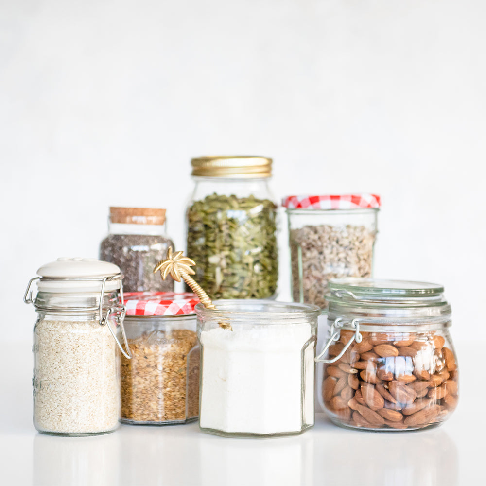 A group of 7 glass jars containing all the seeds and nuts used to make Purition; sesame seeds, flaxseed, coconut, pumpkin seeds, chia seeds, almonds & sunflower kernels.