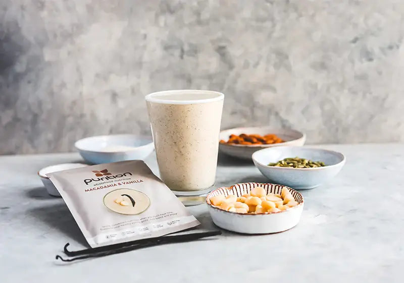Single serving sachet of Purition's Bestselling flavour Macadamia and Vanilla with a shake made up, with pots of seeds of nuts and whole vanilla pods.