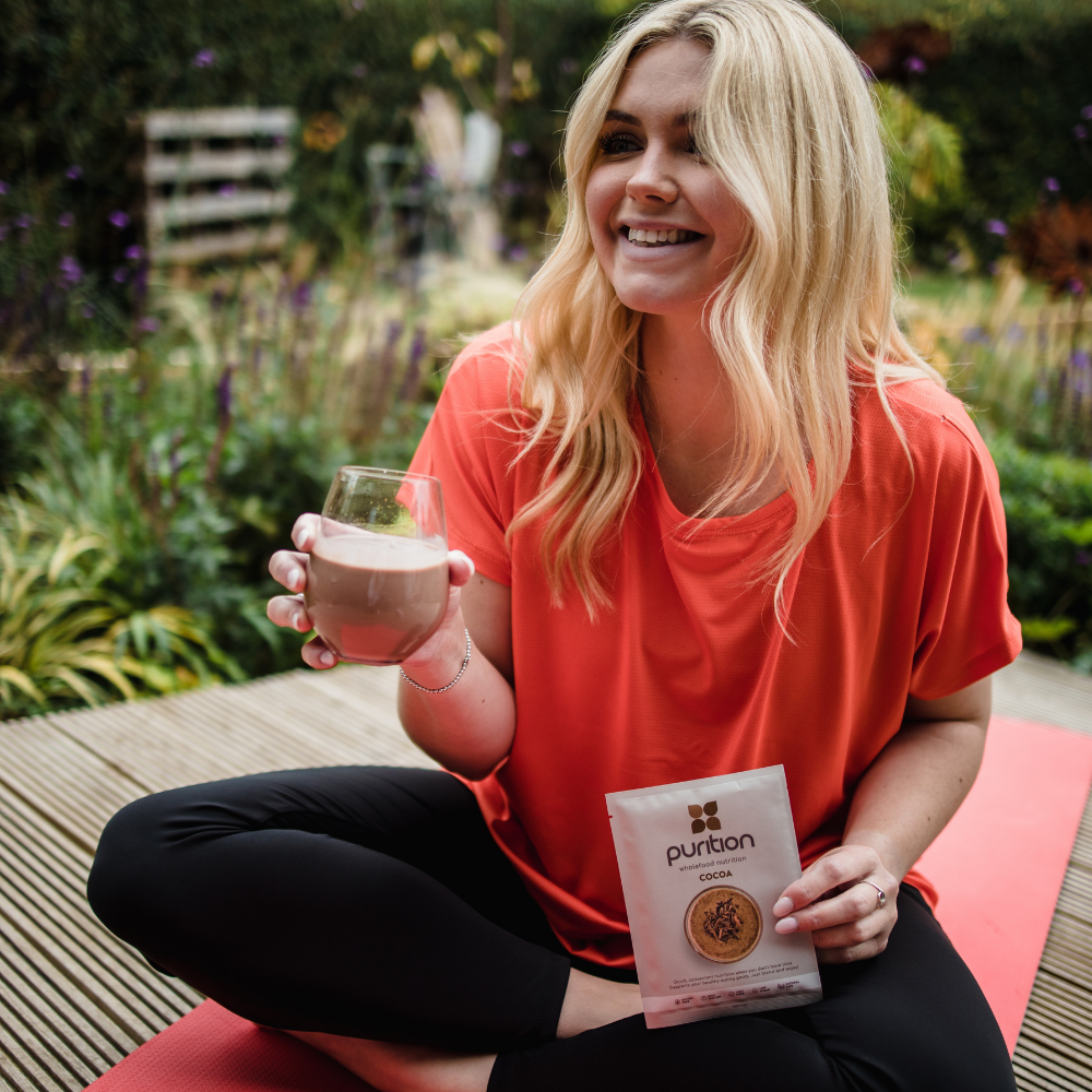 Woman on yoga matt drinking a glass of Purition Chocolate and holding sachet