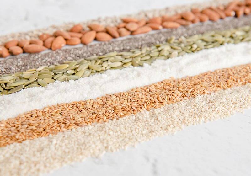 Diagonal narrow stripes of 7 colourful seeds and nuts used to make Purition; sesame seeds, flaxseed, coconut, pumpkin seeds, chia seeds, almonds & sunflower kernels.