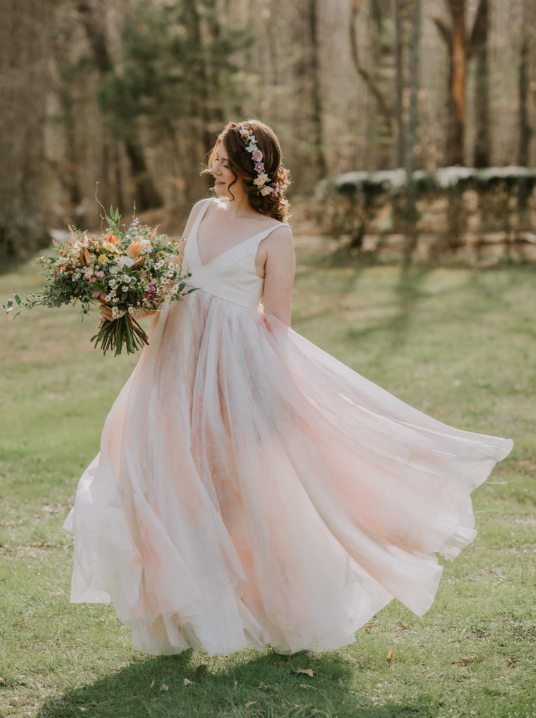 A bride stands on green grass with trees behind her. She has flowers in her hair and holds a boquet. Her white bodice features a deep v-neck and shimmers. Her skirt featuers many layers of translucent silk as it billows and twirls around her legs. Some layers are dyed in shades of peach and soft purple and blue gray.