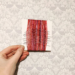 4 inch card vertically wrapped with chunky red and pink yarn, interwoven with single strands of light reflective material.