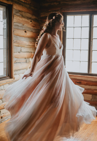 A bride spins in joy in a historic lodge. You can see the log beams of the walls behind her as her dress twirls in the golden light, a smile on her face. Her feet are bare on the hardwood floor. Flowers decorate her hair. 