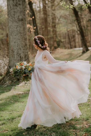 A bride walks through a forest wearing a floaty dress with a many-layered skirt dyed in shades of peach and purple and blue-gray.