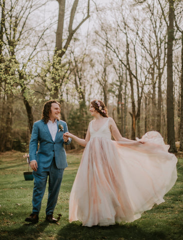 A couple holds hands in front of a copse of trees. He wears a blue suit, she is in a dress with a large skirt that glows in the afternoon light. 