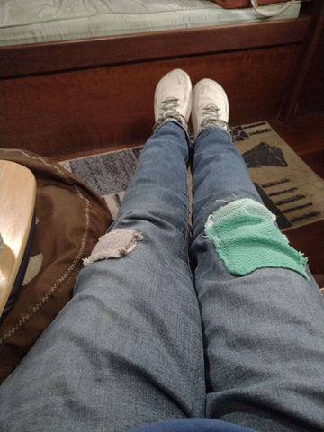 A photo from the view of someone looking at their own legs stretched out. The person is wearing jeans that have big patches on each knee, one is grey and one is bright teal.