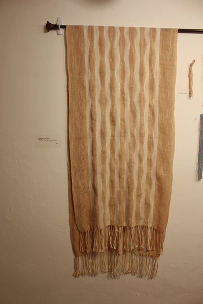 A light-weight and lacy shawl hangs on a rod. It's dominant color is a pink-ish beige, with rippling stripes of creamy white.