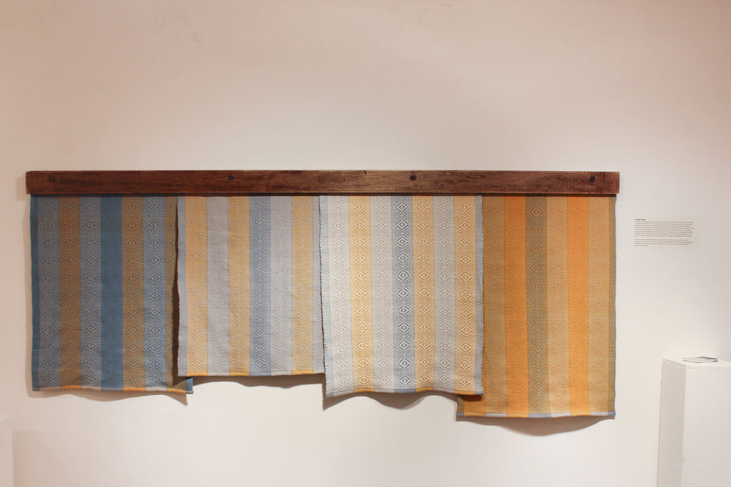 Four kitchen towels hang from a decorative wooden bar. They feature the same stripe design, but have different dominant colors. Left to right: medium blue, pale gray, natural cream, mustard yellow.