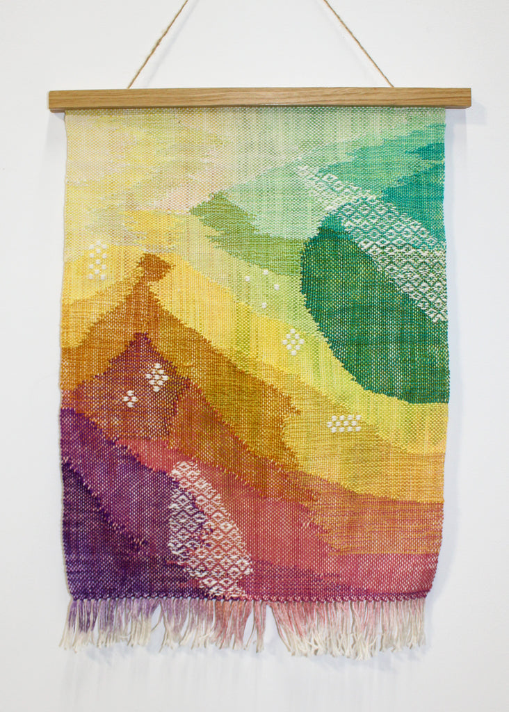 A finished wall hanging on a white wall. The hanging uses bright colors and abstract shapes blended together. The bottom left corner starts with dark purple, and blends into shades of pink, then orange, then yellow, then green in the top right corner.