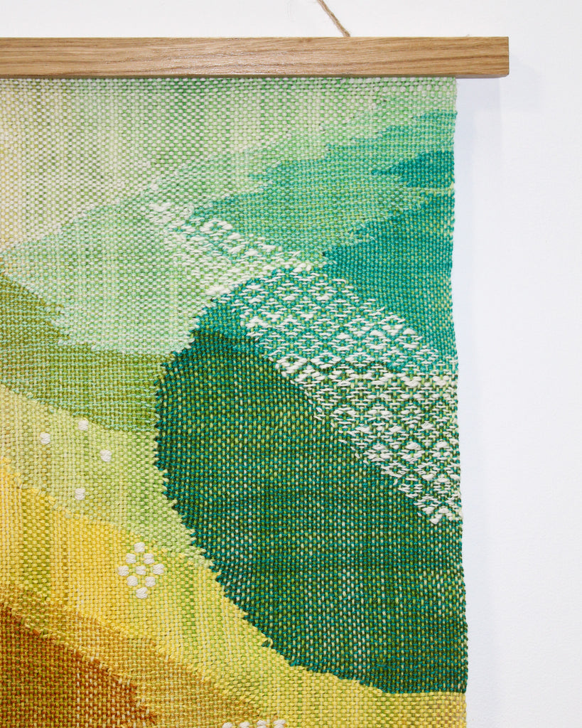 A close up of the top right corner of the hanging. A white textured yarn woven in diamond shapes overlays a background in shades of green.